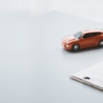 An auto policy holder sits at a table next to a red model car as they sign insurance documents on a clipboard.