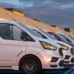 A row of business vehicles parked outside a large commercial warehouse. The vehicles are white work vans.