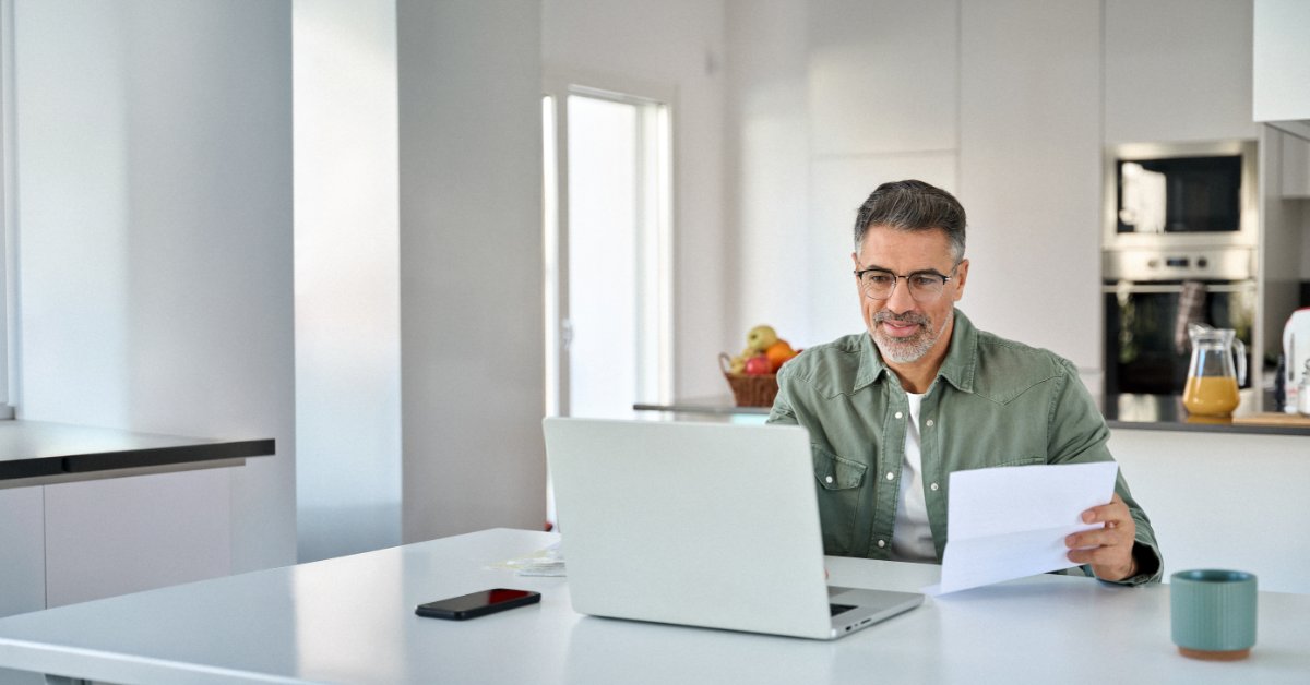 A man wearing glasses sits at a bright kitchen table with a laptop, papers, and a document held in one hand.