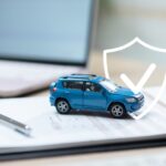 A blue toy car rests on insurance forms with a white shield icon above it. A pen, laptop, and car keys are nearby.