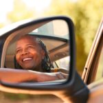 A close-up of a smiling Black woman who looks out the car window from the driver's seat on a sunny day.