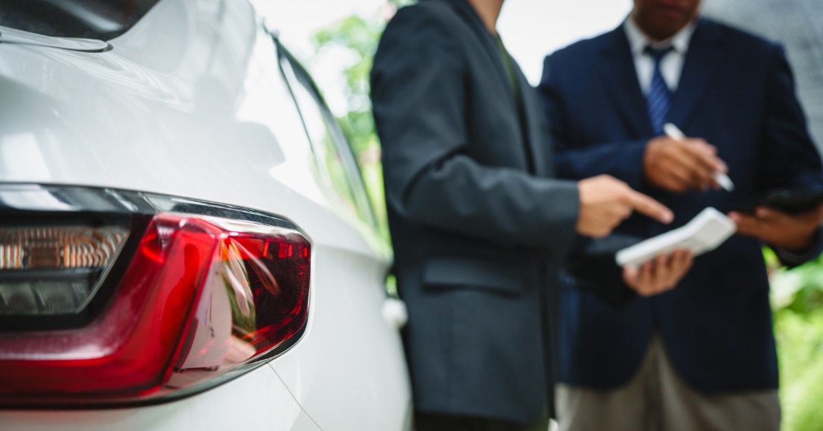 A close-up of two people wearing suits and ties, standing beside a white car in an outdoor parking area.