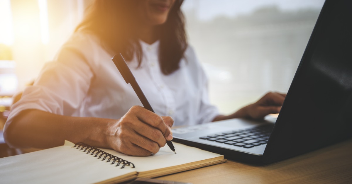 A woman writes in a notebook next to a laptop and phone on her desk, with warm sunlight behind her indoors.