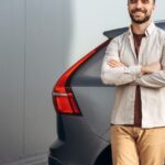 A smiling man stands with his arms crossed beside a parked grey SUV. He wears khaki pants and a long-sleeved shirt.