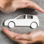 Two hands reaching around a white paper car cutout on a wooden surface with a phone and coffee nearby.