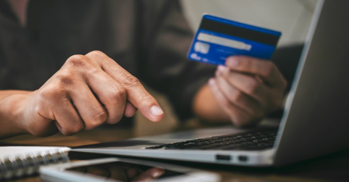 A close-up of a person holding a blue credit card while touching a laptop keyboard, with a phone and notebook nearby.