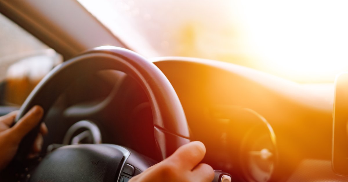 A close-up of a person holding a steering wheel inside a car, with sunlight shining across the dashboard and windshield.