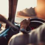 A driver holding a steering wheel inside a car as warm sunlight shines through the windshield and side window.