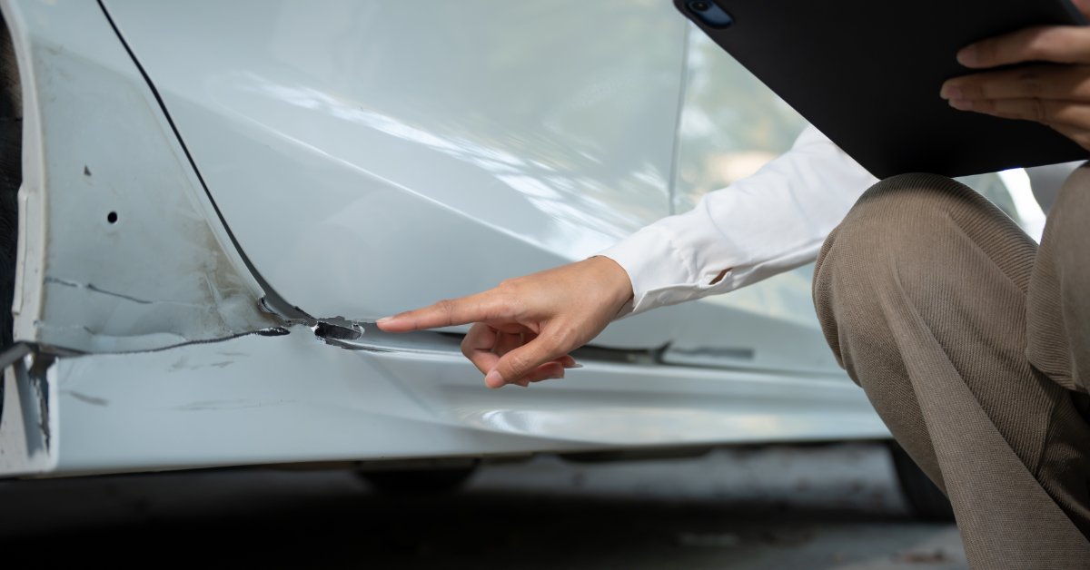 A close-up of a person pointing to scratches and dents on a white car door while holding a black clipboard.