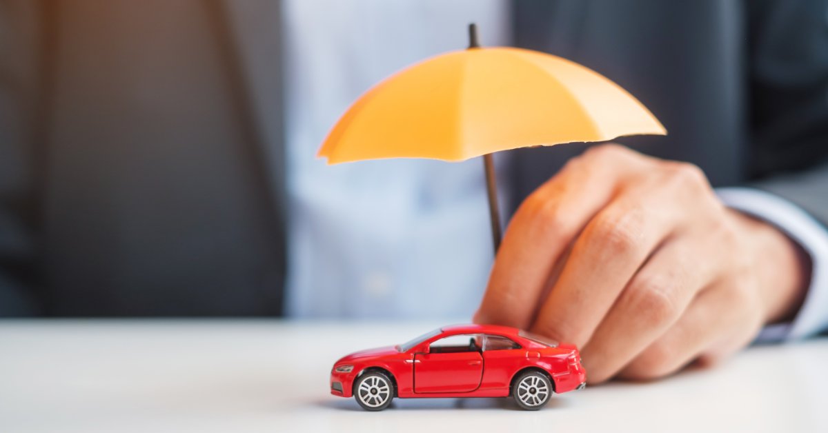 A close-up of a person in a suit holding a small, yellow umbrella over a red toy car on a white table.