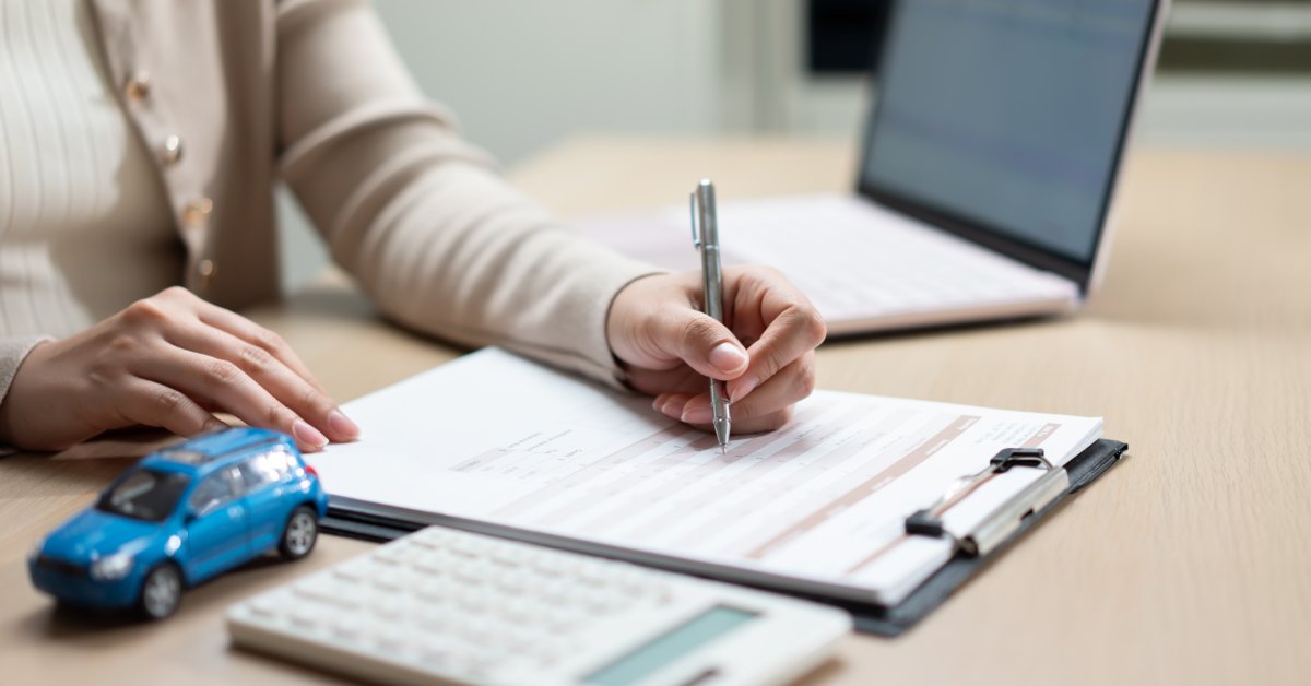 A close-up of a person filling out a car insurance form beside a calculator, pen, and small vehicle model on a desk.