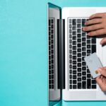 Top view of a person holding a blank credit card while typing on a laptop, sitting on a light-blue surface.