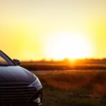A sedan parked beside a rural road with open fields, farm buildings, and a sunset sky in the background.