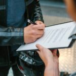 A close-up of a person in a black leather jacket signing a document on a clipboard while holding a motorcycle helmet.