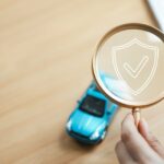 A hand holding a magnifying glass with a white shield and checkmark graphic above a blue toy car on a wooden table.