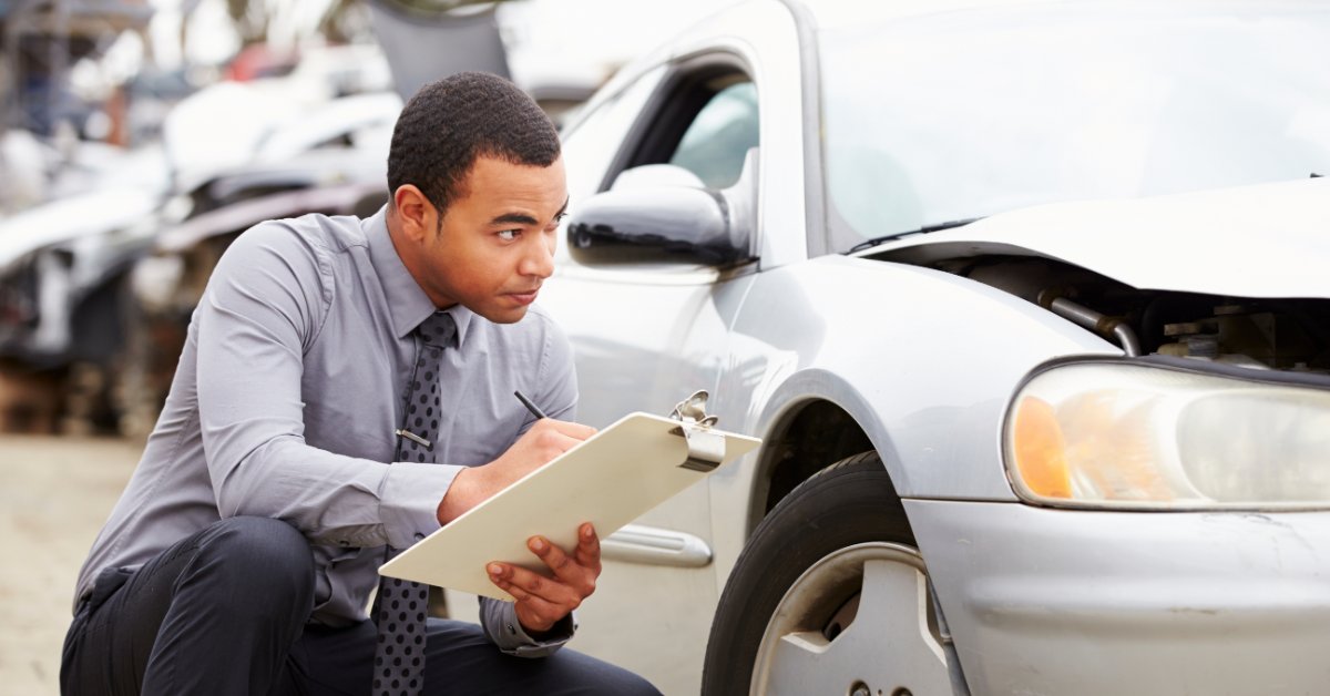A man in a gray shirt and tie crouches beside a damaged silver car. He is writing on a clipboard outdoors.