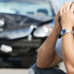 An up-close image of a man wearing a sliver watch with his head in his hands and a wrecked black car in the background.