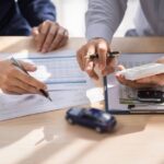 Two people sitting at a table. They are both holding pens over multiple documents, a set of keys, and a toy car.