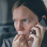 A woman with a nervous expression on her face biting her nails while holding a silver cellphone up to her ear.