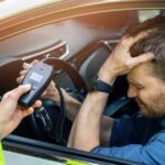 A man in the driver's seat of a vehicle holds his head with his hand. A person stands at the window holding an alcometer.