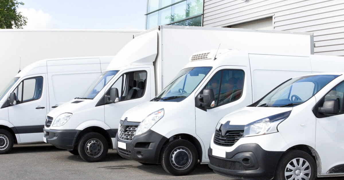 A fleet of five white vans of various sizes parked next to one another in a parking lot. The parking lot is near a building.