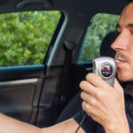 A man sits in the driver's seat of a vehicle while parked and blowing into a breathalyzer test. The man wears a black shirt.