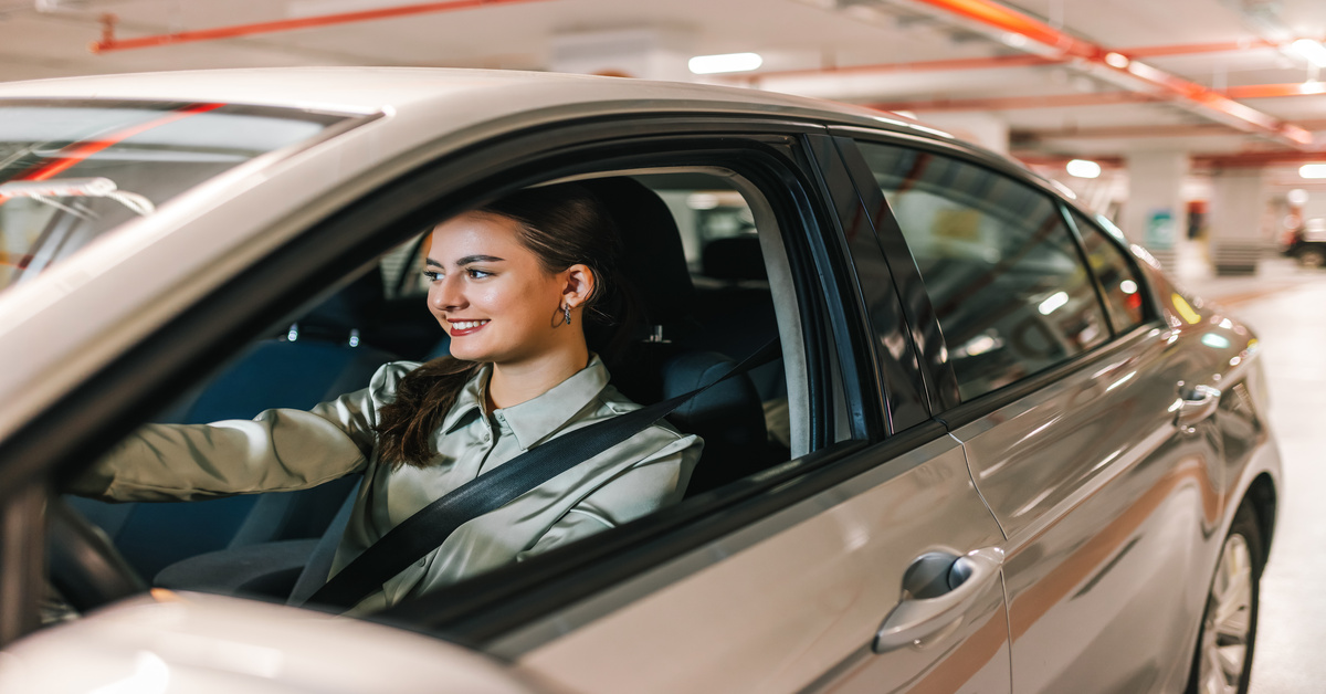 A young woman smiles while looking out the window of her grey car as she drives out of a parking garage.