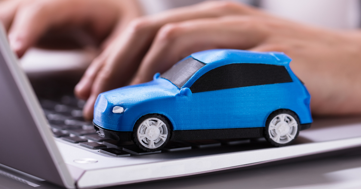 A person sits at a desk while using both hands to type on a laptop. A small blue toy car sits on the laptop.