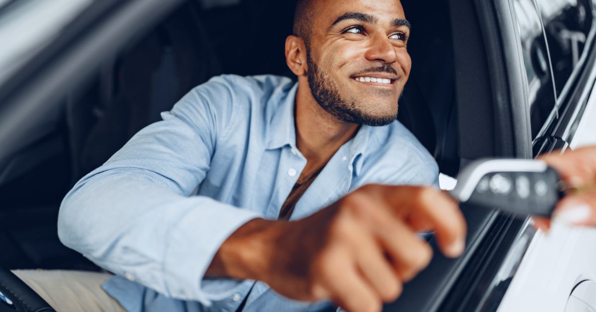 A man in a blue shirt sitting in the driver's seat of a car, smiling while taking the key from someone outside the car.