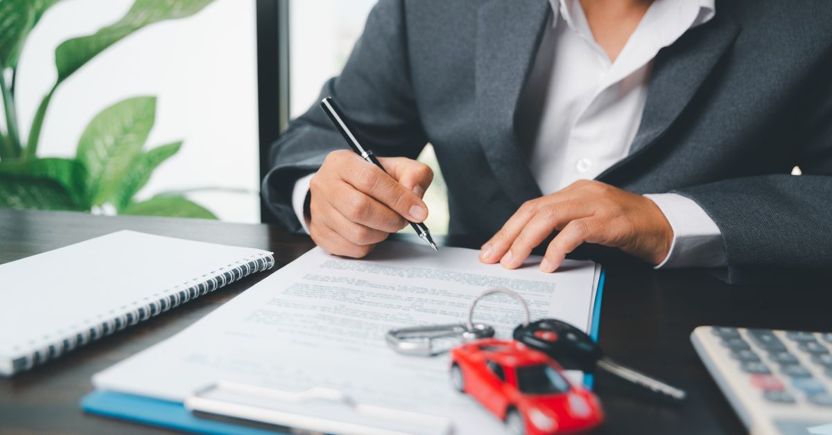 A person in a gray suit sitting at a table while filling out paperwork. A set of keys and a toy car rest on top of the papers.