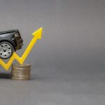A black toy SUV driving up a yellow plastic arrow that sits on top of a stack of gold coins with a gray background.