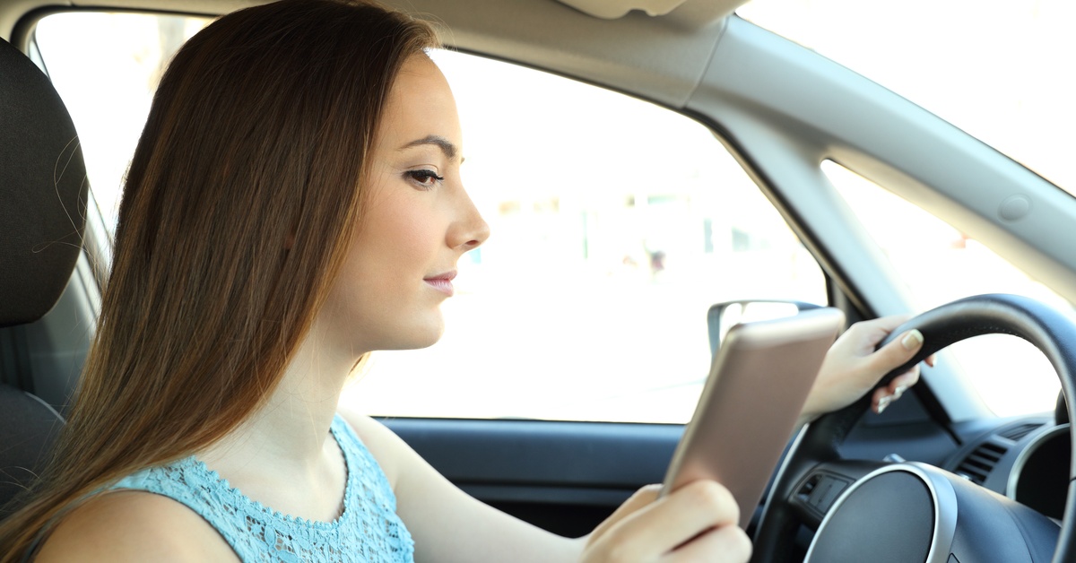 A woman wearing blue sits in the driver's seat of a car, with one hand on the wheel and the other holding a phone.