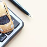 A small gold-colored toy car resting on top of a calculator on a table. A pen sits nearby on the table.
