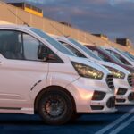 A fleet of generic vans sit parked in a row in a large parking lot. Some vans are white, while others are red or blue.
