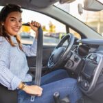 A young woman smiles while putting on her seat belt in the driver's seat of a car. The interior of the car is dark gray.