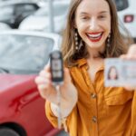 A woman wearing an orange shirt smiles while holding her license and a set of car keys. A car is parked behind her.