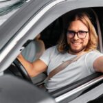 A man wearing glasses sits in the driver's seat of a vehicle. He smiles and dangles the car keys out of the window.