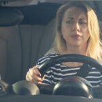 A young woman sits in the driver's seat of a car while holding both hands on the wheel. She watches on with a confused look.