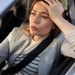 A woman wearing a blazer sits in the driver's seat of a car while leaning her head against her arm on the window.