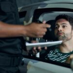 A police officer holding a pen and small notebook stands outside of a parked car with a man sitting inside.