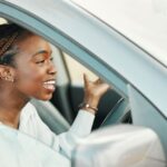 A woman in the driver's seat of a car with the windows down gestures exasperatedly with both hands.