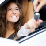 A woman smiles while sitting in the driver's seat of a car and reaches out her hand to accept car keys from someone.