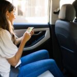 A woman wearing a white shirt sits in the backseat of a car with her seat belt fastened as she looks at a phone.