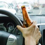 A person sits in the driver's seat of a car, with one hand on the steering wheel and the other holding a beer bottle.