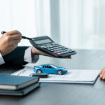 A person wearing a suit holds a pen while pointing toward a calculator. The table contains documents and a toy car.