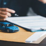 A person sits at a desk while holding a pen to a piece of paper attached to a clipboard. A set of car keys rests nearby.