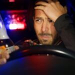 A with a worried expression sits in the driver's seat of a car while holding a glass bottle of alcohol.