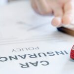 Close-up of a document on a table that reads "Car Insurance Policy." A toy car sits on it while a person signs it.