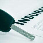 A car key sits on a white surface, next to a document which reads "INSURANCE" at the top of the page in big letters.