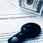 A piece of paper lies on a table, reading "Auto Insurance Policy." A set of car keys and two $100 bills lie on top of it.